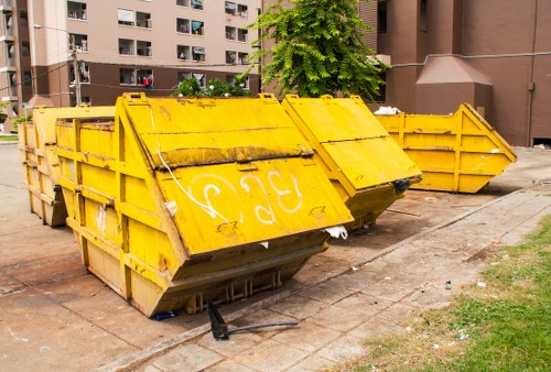 Two large yellow industrial skip containers made of metal with reinforced sides and a weathered finish, positioned on a paved area adjacent to a building. The front skip is tilted forward with its lid partially open, revealing some internal debris. The second skip, aligned behind the first, is upright and closed. Both skips have visible scratches and dirt marks, indicating use for waste disposal. They are situated on a concrete surface with some grass and soil at the edges, typical for commercial waste collection sites in Hatch End. The background features a brown residential or commercial building with multiple balconies and windows, and there are trees providing greenery in the vicinity. The overall scene is well-lit with natural daylight, and the presence of these skips suggests ongoing rubbish removal or waste clearance activities in the area managed by Commercial Waste Hatch End.