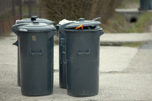 Two dark grey plastic rubbish bins with fitted lids are positioned side by side on a paved driveway. The bin on the right has its lid partially open, revealing some loose waste and an orange object inside. The surface of both bins is slightly textured, with visible scuff marks and minor scratches. The background shows a blurred outdoor setting with trees and a stone bridge or culvert, indicating a residential or suburban area near Hatch End. The bins are aligned on a concrete or tarmac surface, typical for waste collection points, with the surrounding environment suggesting a tidy external space suitable for domestic rubbish management. This image relates to rubbish removal services offered by Commercial Waste Hatch End, serving local areas including HA5 postcode neighborhood, emphasizing responsible waste disposal and collection.