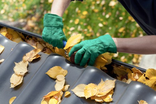 A person wearing green gardening gloves is collecting fallen yellow leaves onto a dark grey plastic gutter with a slightly corrugated surface. The individual is using their hands to scoop leaves, some of which are perched on the gutter's curved ridges, indicating a process of clearing outdoor debris. The background appears to feature a garden or outdoor space with blurred greenery and scattered leaves, suggesting an autumn setting. The gutter is situated on an outdoor area, likely a residential property, and the scene relates to routine rubbish removal or garden maintenance, services that Commercial Waste Hatch End provides in the local area near HA5 postal code. The focus is on the detailed, textured leaves and the gloved hands actively involved in tidying an outdoor space, exemplifying typical waste management tasks related to garden clearance and rubbish removal.