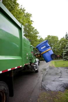 A green waste collection truck from Commercial Waste Hatch End is parked on a residential street in Hatch End, with its rear section visible. The truck is equipped for rubbish removal, featuring a smooth, metallic green finish and a large side panel. A person, only partially visible, is lifting a blue wheeled bin towards the truck’s open rear loading area, which is designed for efficient collection of household or commercial waste. The blue bin, made of plastic, has a lid and is positioned at an angle, suggesting it is being emptied or loaded. The scene occurs on a paved road with a slight curve, bordered by a grassy verge and a concrete driveway with an asphalt surface. In the background, tall leafy trees and a well-maintained suburban environment are visible, with overcast lighting indicating a cloudy day. This image captures a typical rubbish collection process in Hatch End, aligning with local waste management services offered by Commercial Waste Hatch End in the area with postcode HA5, emphasizing their rubbish removal capabilities.