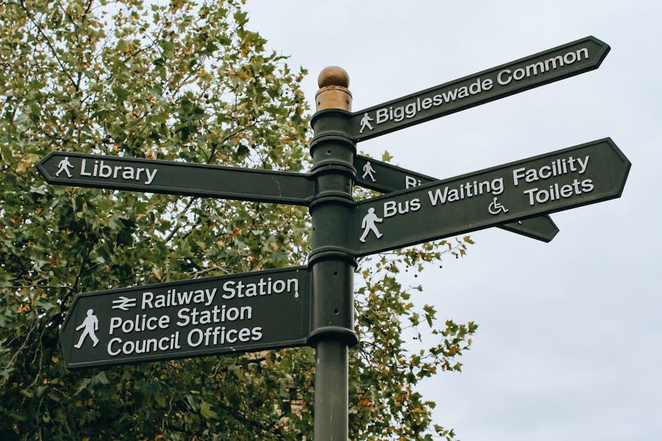 A black directional street signpost standing amidst green leafy trees, with multiple rectangular signs pointing in different directions. The signs feature white text and pedestrian icons, indicating l
